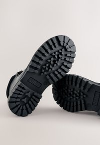 Black rugged boots with thick, textured rubber soles featuring aggressive treads, and embossed “NEXT” logo on the side of each sole.