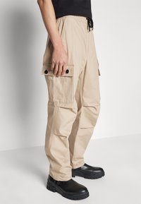 Beige cargobroek met een elastische tailleband en trekkoord. Voorzien van twee zijvakjes met zwarte knopen en een relaxed fit. Draagt met zwarte laarzen.