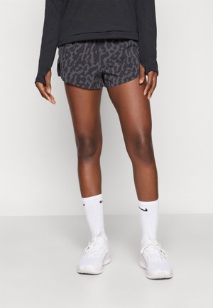 Osoba ubrana w czarne i szaro wzorzyste sportowe spodenki, czarną bluzę z długim rękawem, białe skarpetki Nike do kostek oraz białe trampki, stojąca na szarej podłodze.