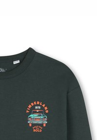 T-shirt Timberland vert foncé avec un graphisme orange et turquoise d'une voiture et le texte "Timberland 1973 Conçu pour les audacieux" sur la poitrine gauche.