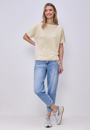 Donna sorridente con capelli biondi che indossa una maglietta beige a maniche corte, jeans azzurri e scarpe da ginnastica grigie e blu, in piedi contro un muro chiaro.