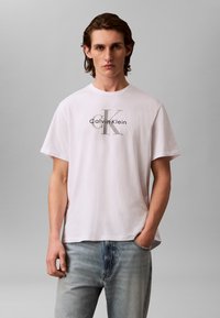 Hvit bomulls-T-skjorte med en grafisk logo i svart og gr ått midt på, kombinert med lyseblå denimjeans.