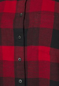 Camicia flanella a quadri rossi e neri con chiusura a bottoni, taschino sul petto e tessuto testurizzato. I bottoni sono scuri e circolari.