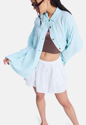Kobieta ubrana w za dużą, jasnoniebieską koszulę w paski, na brązowym crop topie i białej spódnicy, pozująca na tle jednolitego białego tła.