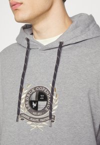 Sudadera con capucha gris y cordones, con un logo bordado que incluye una corona floral y detalles de texto. Tejido suave con un ajuste relajado.
