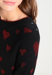 Zwarte trui met rode glitterharten, ronde halslijn met een contrasterende rode rand en zwarte knoopdetails op de schouder.