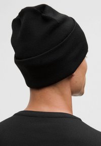 Czarna dzianinowa czapka z taperowanym, zaokrąglonym wierzchem i podwiniętym mankietem. Tkanina wydaje się miękka i elastyczna, współgrając z czarną teksturowaną koszulą poniżej.