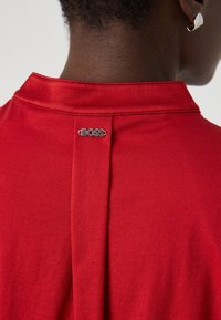 Camicia rossa con colletto e piega sul retro, contraddistinta da un logo "BOSS" in argento. Il materiale ha una texture liscia.