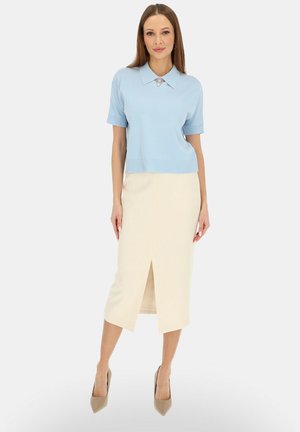 Kobieta w jasnoniebieskiej bluzce z krótkim rękawem i kołnierzykiem oraz kremowej midi spódnicy z przednim rozcięciem, stojąca na tle jednolitego tła.