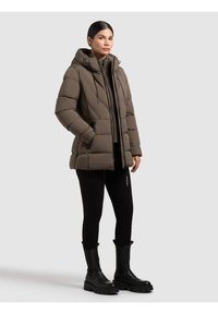 Pufjakke i afdæmpet brun med hætte, lynlåslommer og kviltet design. Kombineret med sorte leggings og kraftige sorte støvler.