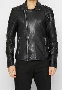 Mężczyzna w czarnej skórzanej kurtce motocyklowej z asymetrycznym zapięciem na suwak i kieszeniami na zamek po bokach, zestawionej z czarnymi spodniami.