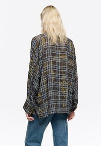 Camisa de cuadros marrón semitranslúcida con patrones en amarillo y azul, corte relajado, mangas largas y puños abotonados. Tejido texturizado con una silueta fluida.
