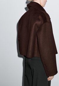 Krótka brązowa wełniana kurtka z wysokim kołnierzem i luźnym krojem, o skróconym fasonie i lekkim rozszerzeniu u dołu.