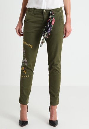 Pantalon slim vert olive avec broderies florales et inscription « Eighty-One California », associé à un foulard fleuri noué à la taille et des talons hauts noirs.