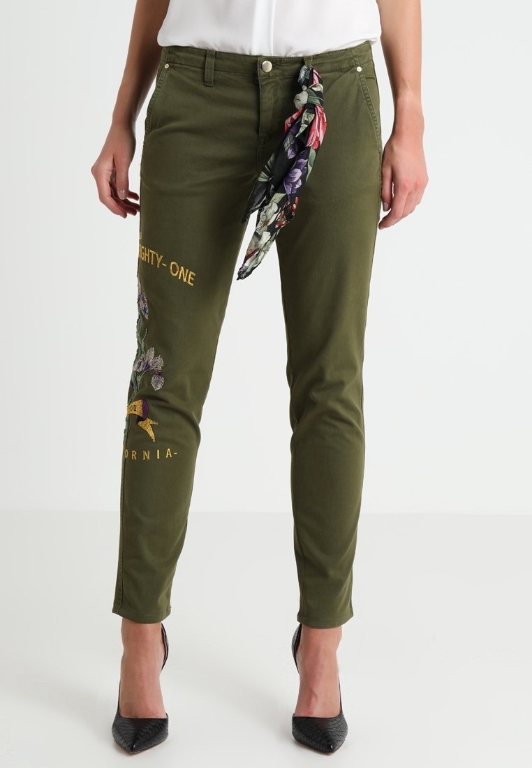 Pantalon slim vert olive avec broderies florales et inscription « Eighty-One California », associé à un foulard fleuri noué à la taille et des talons hauts noirs.