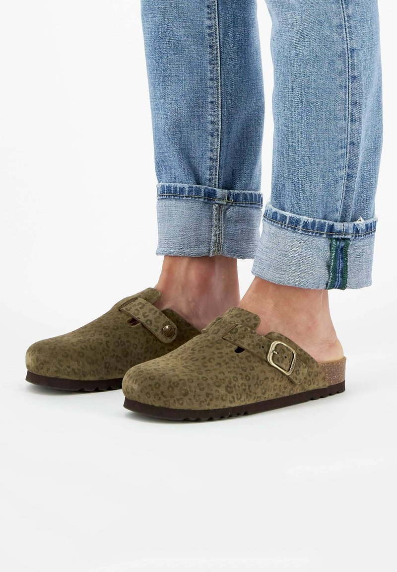 Füße in olivgrünen, gepunkteten Clogs mit Schnallen, kombiniert mit hochgekrempelten, hellblauen Jeans auf einem weißen Hintergrund.