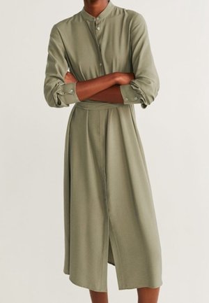Osoba ubrana w oliwkowozieloną sukienkę midi na długi rękaw, zapinaną na guziki, z kołnierzykiem stójką, stojąca z założonymi rękami.