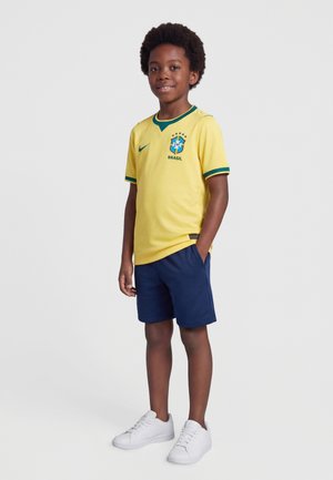 Dreng står med hænderne i lommerne iført gul Brasilien fodboldtrøje, marineblå shorts og hvide sneakers på ensfarvet baggrund.