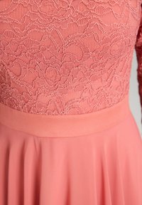 Robe rose avec un corsage en dentelle florale et une jupe fluide. Le tissu est lisse, la taille est cintrée et les manches sont longues.