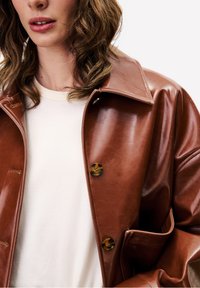 Vrouw met golvend bruin haar draagt een glanzend, bruin leren jasje over een witte top, tegen een effen lichte achtergrond.