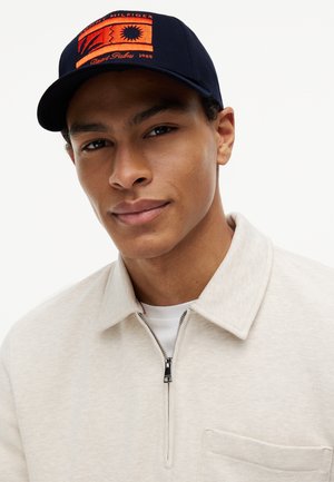Young man wearing navy cap with orange patch and beige zip-up jacket over white shirt, looking at camera with slight smile.