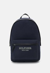 Zaino in tessuto navy con forma curva, tasca frontale con zip, dettagli neri e logo bianco "HILFIGER NEW YORK". Texture liscia, design minimal.