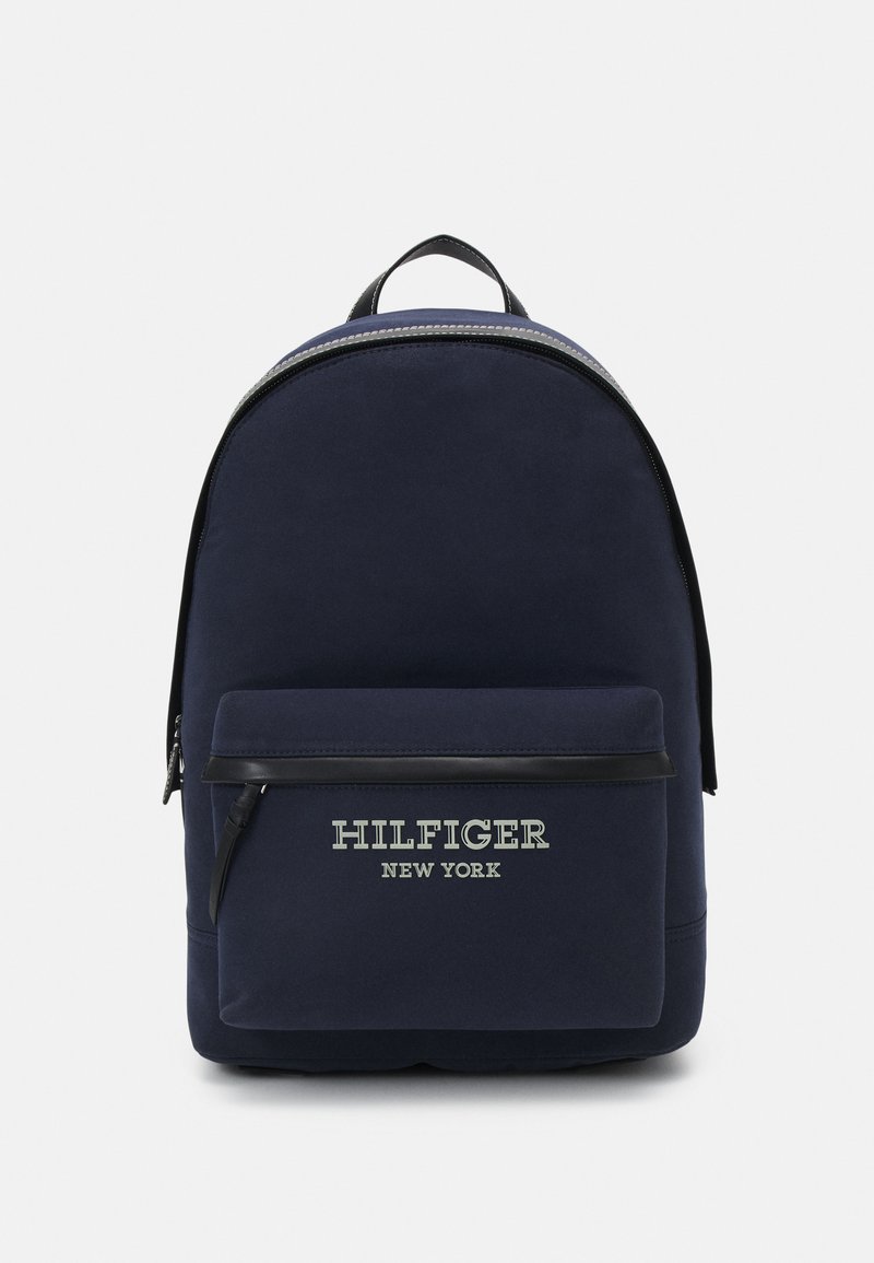 Zaino in tessuto navy con forma curva, tasca frontale con zip, dettagli neri e logo bianco "HILFIGER NEW YORK". Texture liscia, design minimal.