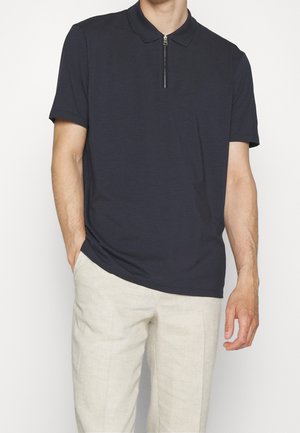 Homme portant un polo noir à manches courtes avec une fermeture éclair à l'avant et un pantalon beige clair, debout avec une main dans la poche.