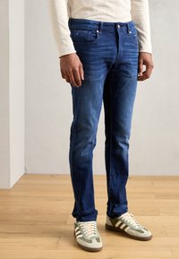 Schmal geschnittene Jeans aus Denim in Dunkelblau, mit fünf Taschen, einem Knopfverschluss und sanften Verblassungen. Kombiniert mit grauen und grünen Sneakers.