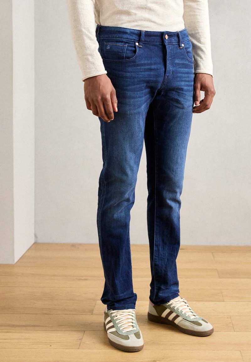 Sötétkék, slim fit farmernadrág, öt zsebbel, gombos cipzárral és enyhe kopásokkal. Szürke és zöld sportcipővel párosítva.