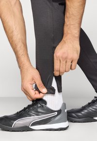 Μαύρο αθλητικό παντελόνι με υφή υφάσματος, που διαθέτει φερμουάρ κοντά στον αστράγαλο. Το χέρι ενός ατόμου ρυθμίζει το φερμουάρ πάνω από λευκές κάλτσες και μαύρα παπούτσια.