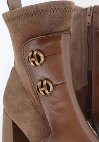 Stivale alla caviglia in pelle marrone e suede caratterizzato da accenti di hardware circolari dorati, dettagli testurizzati e un tacco spesso. Chiusura con zip laterale.