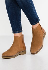 Bottines en suède marron avec des panneaux latéraux élastiques, des coutures décoratives et un petit talon carré, associées à un jean bleu foncé.