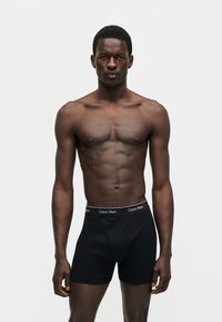 Μυώδης άνδρας στέκεται όρθιος φορώντας μαύρα boxer briefs Calvin Klein σε απλό λευκό φόντο.