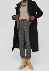 Long manteau noir, pull beige, pantalon gris à carreaux, bottines camel à talon carré, avec un cordon autour du cou et des ourlets retroussés.