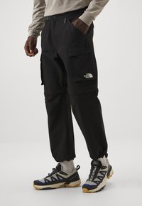 Czarne spodnie cargo z wieloma kieszeniami, elastycznymi mankietami i naszywką z logo; w zestawie z ciemnoniebieskimi i szarymi butami trekkingowymi.