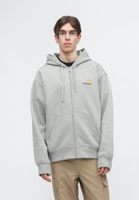 Siva zip-up dukserica od flisa, s prednjim džepom, vezicama i Carhartt logom na prsima. Dizajn opuštenog kroja.