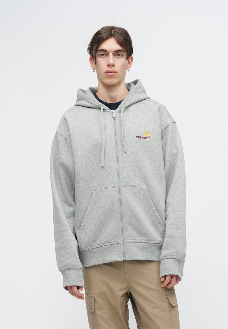 Grauer Reißverschluss-Hoodie aus Fleece-Material, mit einer Fronttasche, Kordelzügen und Carhartt-Logo auf der Brust. Entspannte Passform.