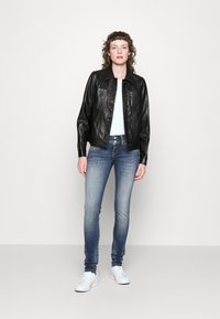 Chaqueta de cuero negra con un diseño con cuello, cierre de botones y dos bolsillos en el pecho, combinada con jeans ajustados azules y zapatillas blancas.