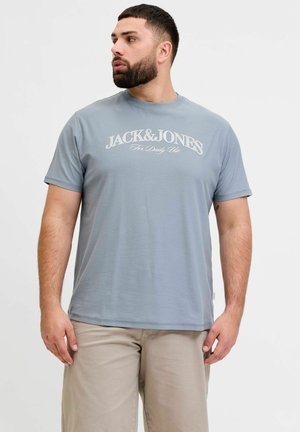 Mężczyzna z brodą, ubrany w jasnoniebieską koszulkę "Jack & Jones" i beżowe spodnie, patrzący w lewo na jednolitym białym tle.