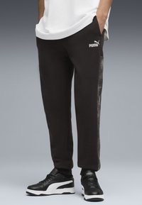 Pantaloni sport negri cu un design ajustat, având un panou lateral texturat și un logo Puma alb. Împreună cu adidași negri și albi.