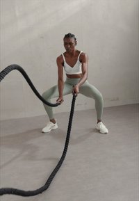 Corda da battaglia nera tenuta da una persona in un reggiseno sportivo bianco e leggings verde chiaro, su un pavimento grigio chiaro contro un muro semplice.