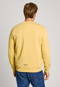 Felpa gialla con collo a giro, polsini e orlo a costine. Presenta un piccolo logo sul retro vicino al fondo. Indossata con jeans blu.