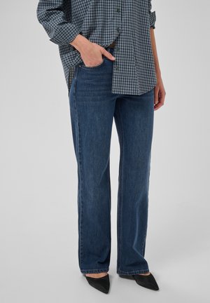 Personne portant un jean droit bleu, des chaussures noires pointues et une chemise à carreaux rentrée d'un côté et pas de l'autre, avec une main dans la poche.