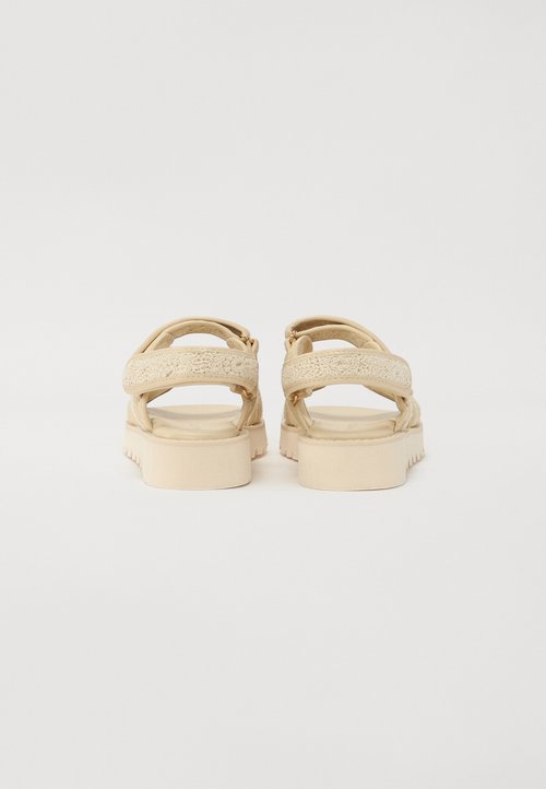 Sandalias beige con dos correas cruzadas, acentos texturizados, hebillas ajustables y una suela robusta y acolchada. Perfil plano y sin patrones notables.