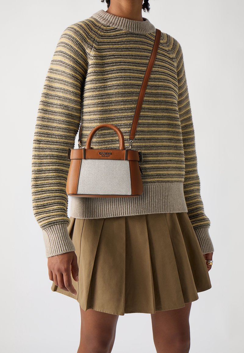 Personne portant un pull rayé beige et gris et une jupe plissée kaki, portant un petit sac à main marron et blanc avec une bandoulière.