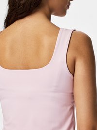 Γυναίκα με μέτριο τόνο δέρματος που φοράει απαλό ροζ αμάνικο τοπ, απεικονιζόμενη από πίσω και πλάγια, με φόντο ένα απλό λευκό σκηνικό.