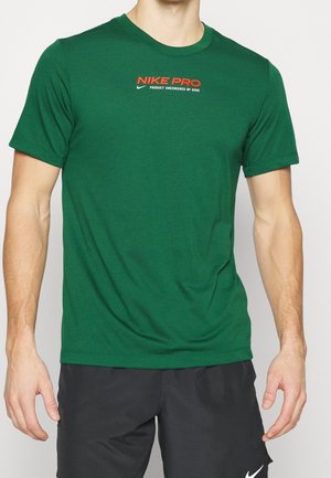 Homme portant un t-shirt Nike Pro vert avec un logo rouge et un short Nike noir, debout devant un fond uni.