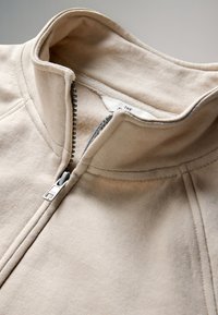 Beige Reißverschlussjacke mit hohem Kragen, sichtbarer Metallreißverschluss teilweise geöffnet, zeigt weiches Innenfutter und abgesteppten Nähten.