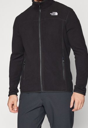 Czarna polarowa kurtka zapinana na zamek z logo The North Face na piersi, noszona przez osobę w ciemnych spodniach, ręce rozluźnione wzdłuż boków.